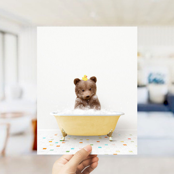 A hand holds up a framed image of a small brown bear sitting in a yellow bathtub filled with bubbles, with a rubber duck perched on its head.