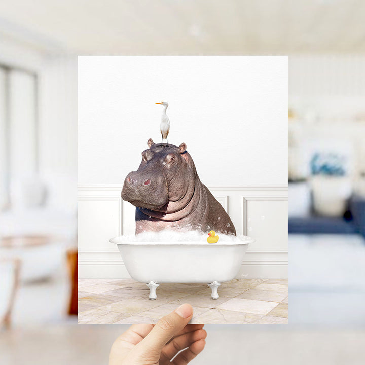 A hand holds up a framed image depicting a hippopotamus sitting in a bathtub, with a seagull perched on its head.