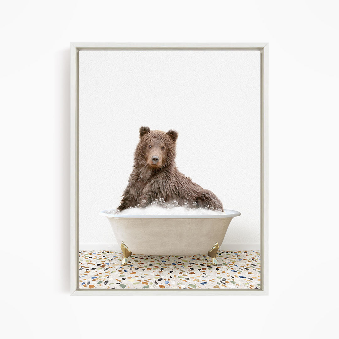 A brown bear sitting in a white bathtub filled with bubbles, looking directly at the camera.