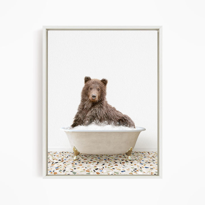 A brown bear sitting in a white bathtub filled with bubbles, looking directly at the camera.