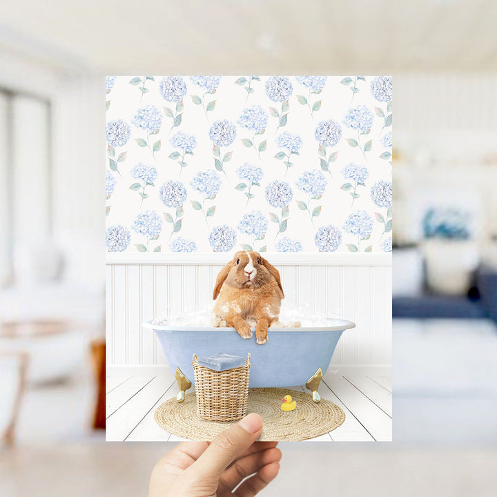A hand holds up a photograph of a brown rabbit sitting in a blue bathtub filled with water, surrounded by a floral patterned wall.
