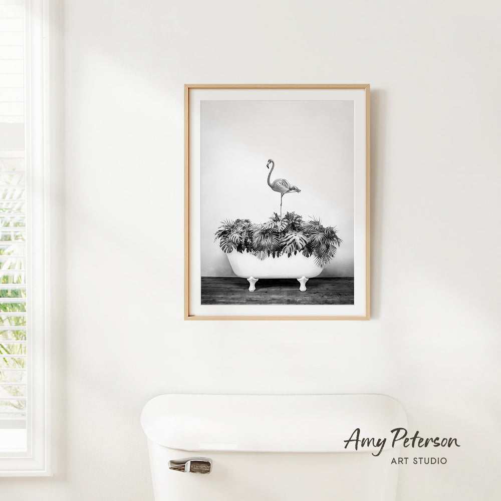 A framed black and white photograph of a flamingo standing on a bathtub filled with plants is hanging on a white wall.