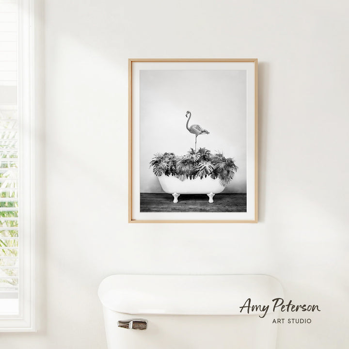 A framed black and white photograph of a flamingo standing on a bathtub filled with plants is hanging on a white wall.