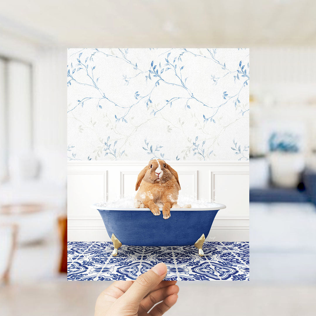 A hand holds up a framed image of a brown rabbit sitting in a blue bathtub filled with water, surrounded by a blue and white patterned floor.