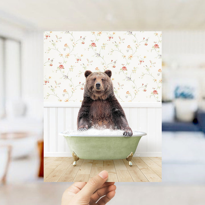 A hand holds up a framed image of a brown bear sitting in a green bathtub filled with bubbles, against a floral wallpaper backdrop.