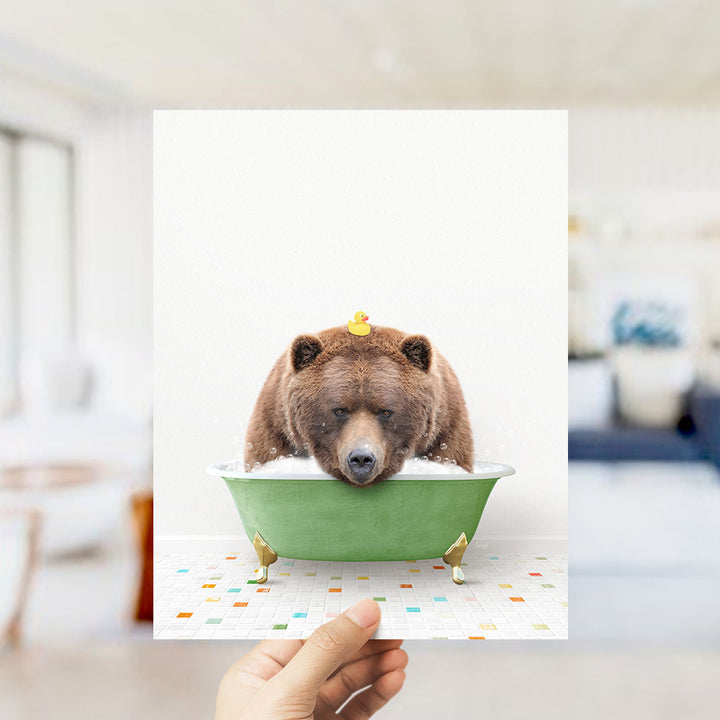 A hand holds up a framed image of a brown bear sitting in a green bathtub, with a yellow rubber duck floating on its head.