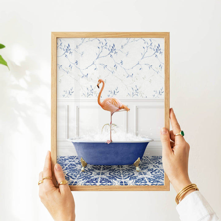 A person's hands are holding a framed picture of a pink flamingo sitting in a blue bathtub, with a blue and white floral pattern on the wall behind the flamingo.