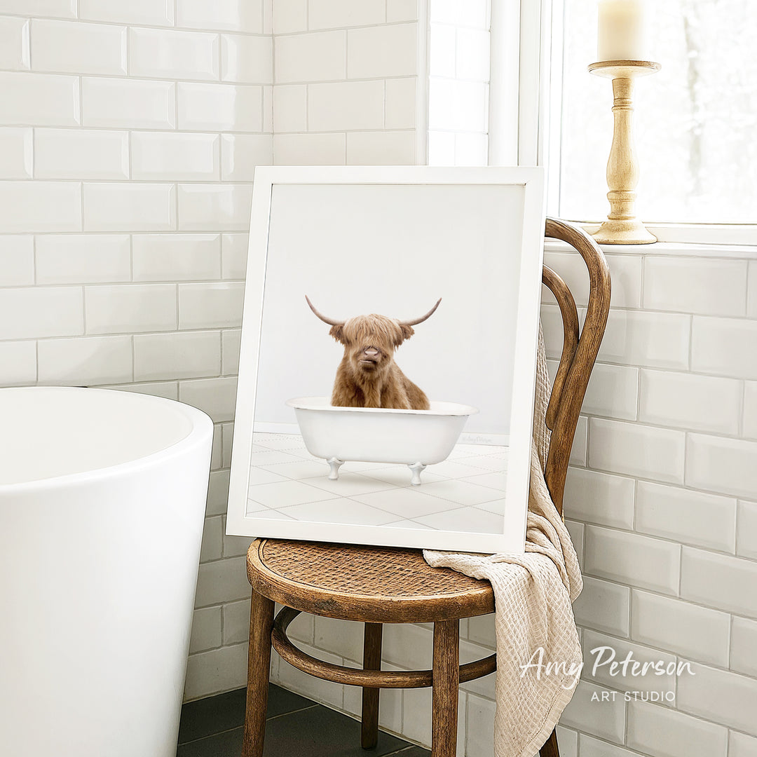 A framed image of a brown cow sitting in a white bathtub, placed on a wooden chair in a bathroom setting.