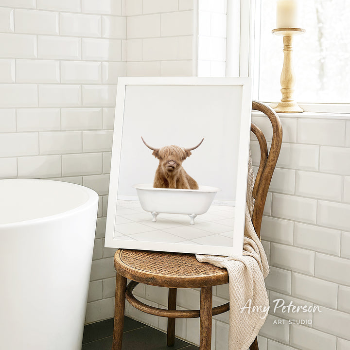 A framed image of a brown cow sitting in a white bathtub, placed on a wooden chair in a bathroom setting.