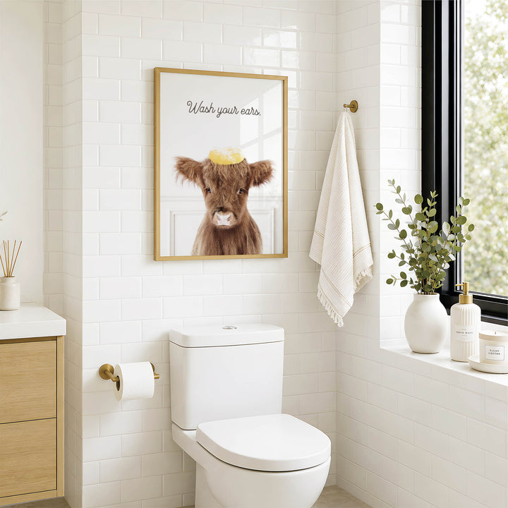 A bathroom with a white toilet, a framed picture of a cow with a yellow object on its head, and a towel hanging on a rack.