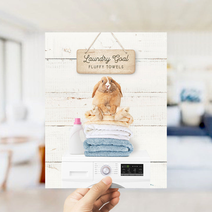 A hand holding a sign that says "Laundry Goal Fluffy Towels" with a picture of a fluffy towel and a rabbit on top of it.