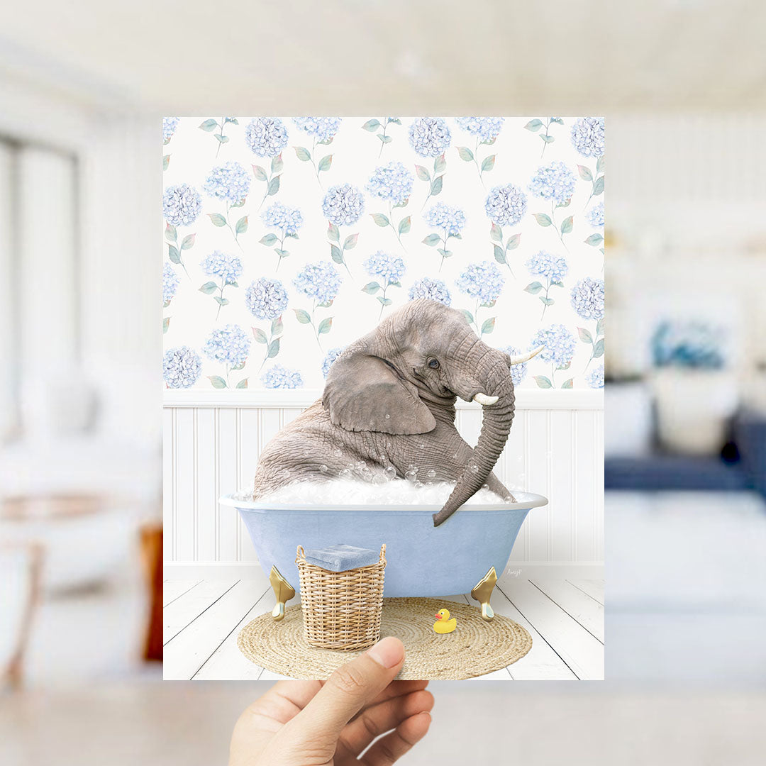 A hand holds up a framed image depicting a gray elephant sitting in a blue bathtub filled with bubbles, surrounded by a wicker basket and a yellow rubber duck.