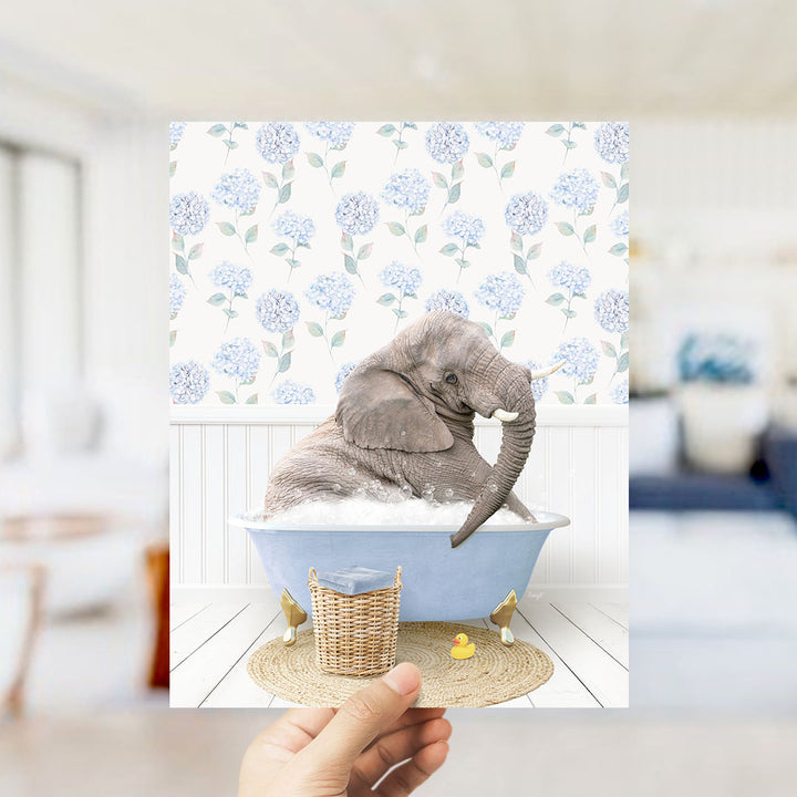 A hand holds up a framed image depicting a gray elephant sitting in a blue bathtub filled with bubbles, surrounded by a wicker basket and a yellow rubber duck.