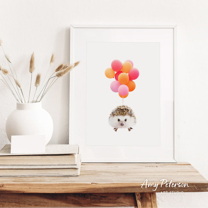 a white vase with dried reeds, a stack of books, and a framed print of a hedgehog with colorful balloons floating above it.