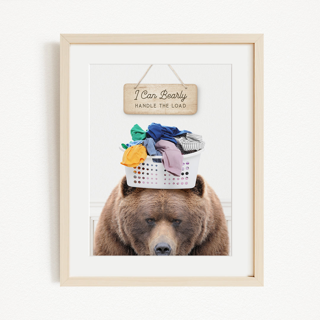 A framed image of a brown bear sitting on top of a laundry basket filled with clothes, with a wooden sign above the bear that says "I Can Bearly Handle the Load".