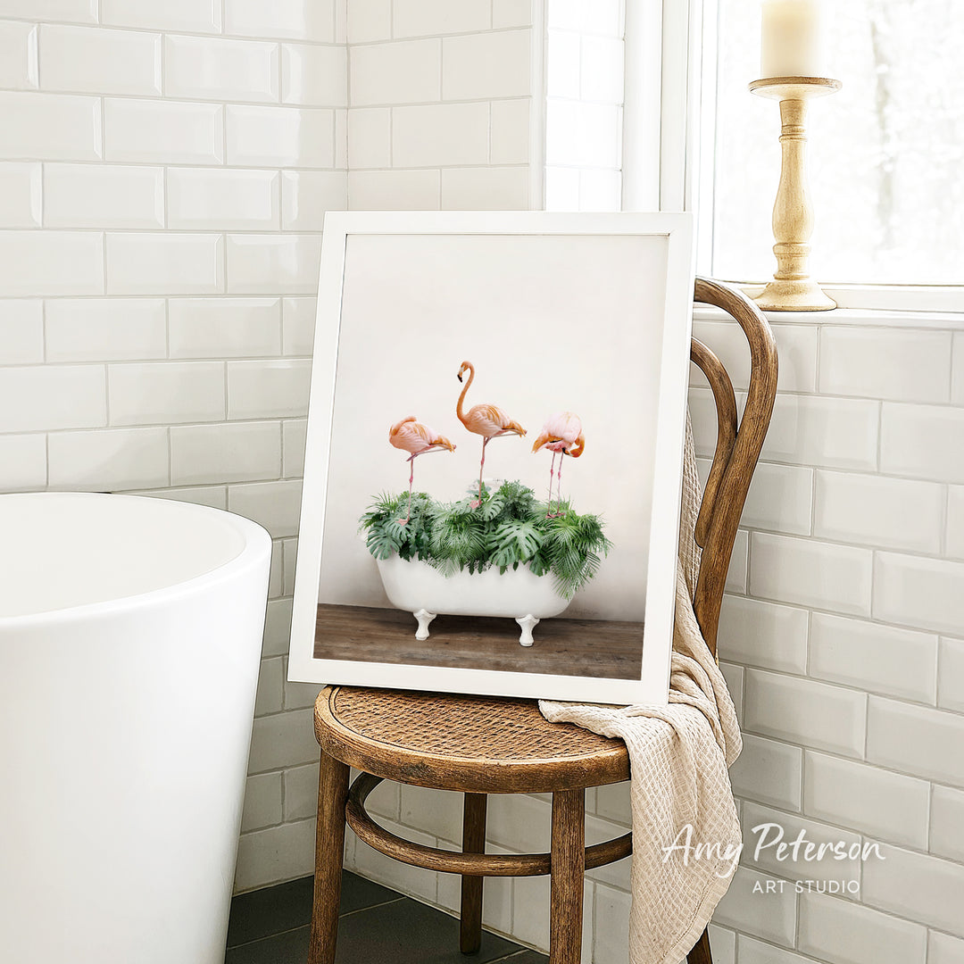 A framed image of three pink flamingos standing in a white bathtub filled with greenery, displayed on a wooden chair in a bathroom setting.