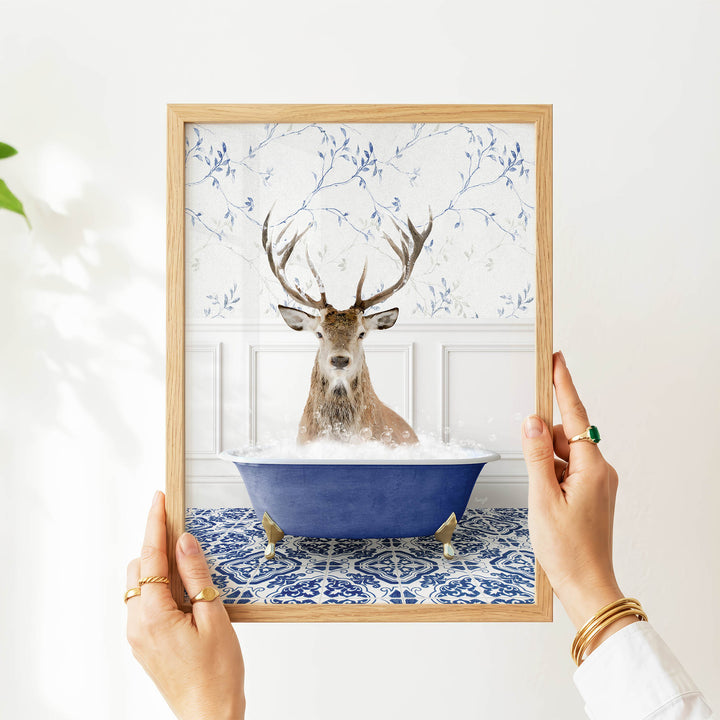 A person's hands are holding a framed picture of a deer sitting in a blue bathtub filled with water, surrounded by a blue and white floral pattern.