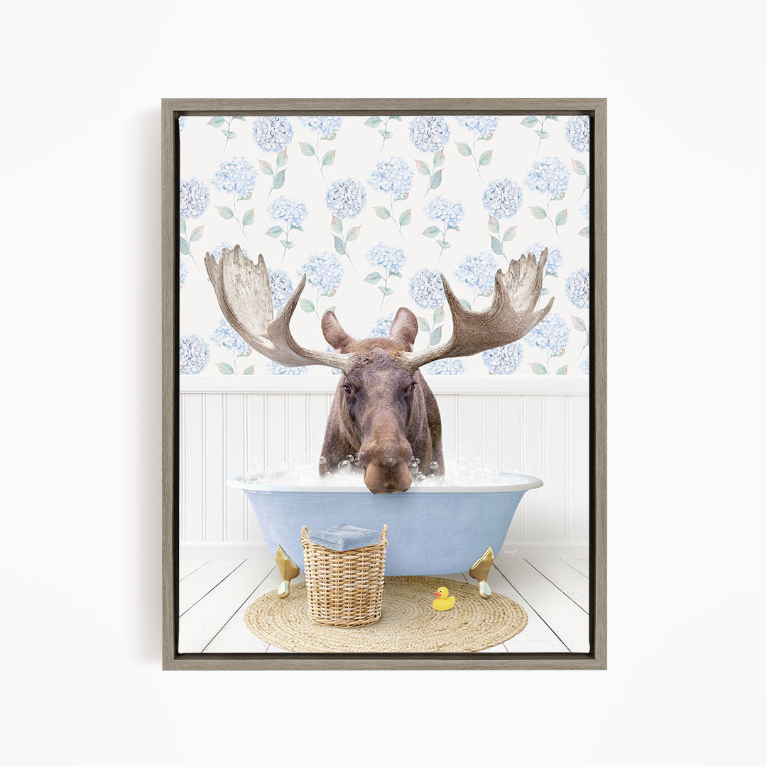 A moose with large antlers is sitting in a blue bathtub filled with water, surrounded by a wicker basket and yellow rubber ducks.