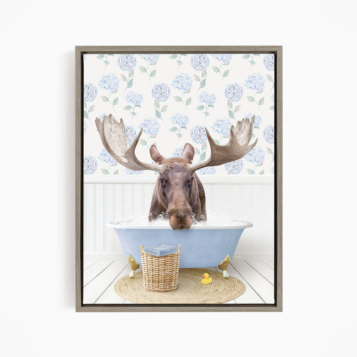 A moose with large antlers is sitting in a blue bathtub filled with water, surrounded by a wicker basket and yellow rubber ducks.