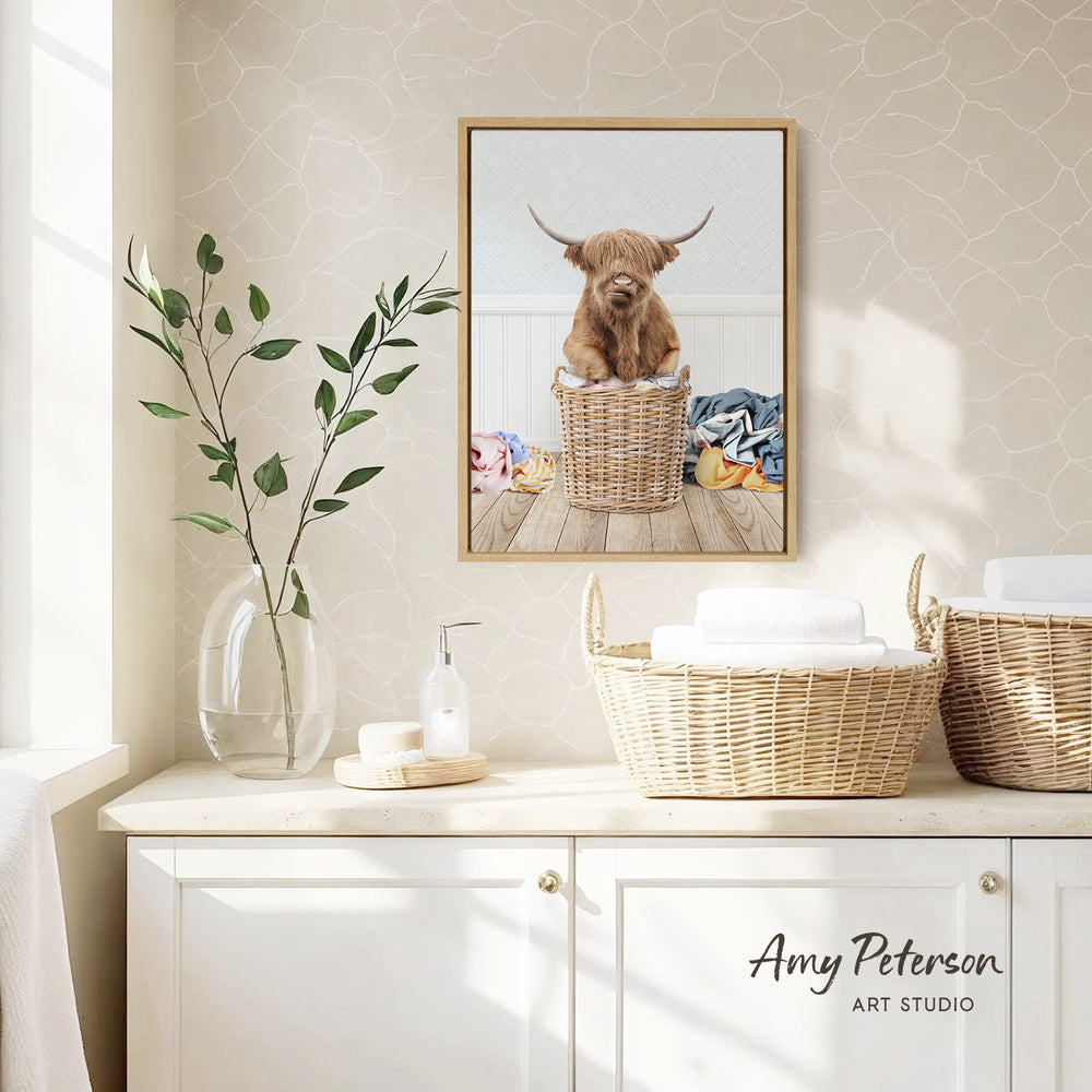 a framed picture of a Highland cow sitting in a wicker basket, placed on a white cabinet in a bathroom setting.