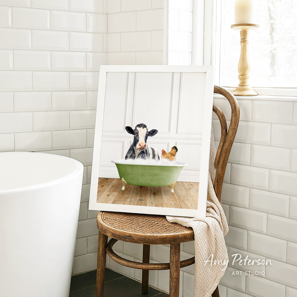 A framed image of a cow sitting in a bathtub, with a chicken nearby, is displayed on a wooden chair in a bathroom setting.