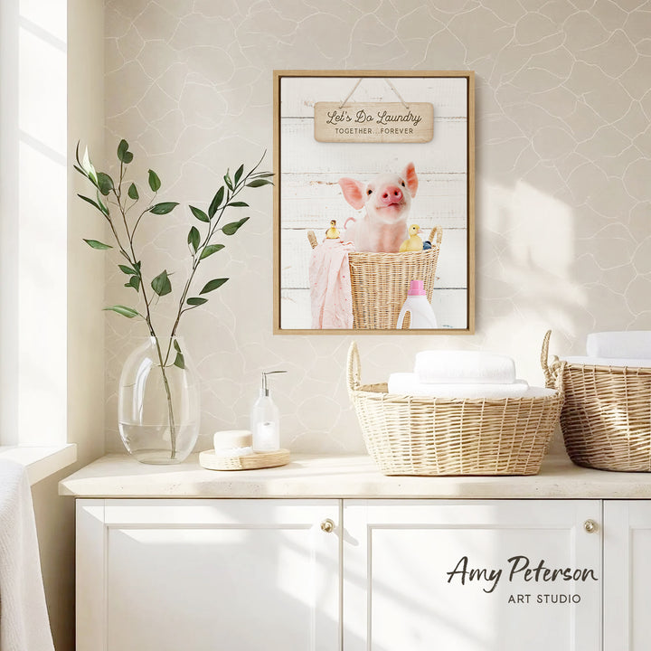 a bathroom scene with a framed picture of a pig sitting in a basket, a vase with green leaves, and a basket with towels.
