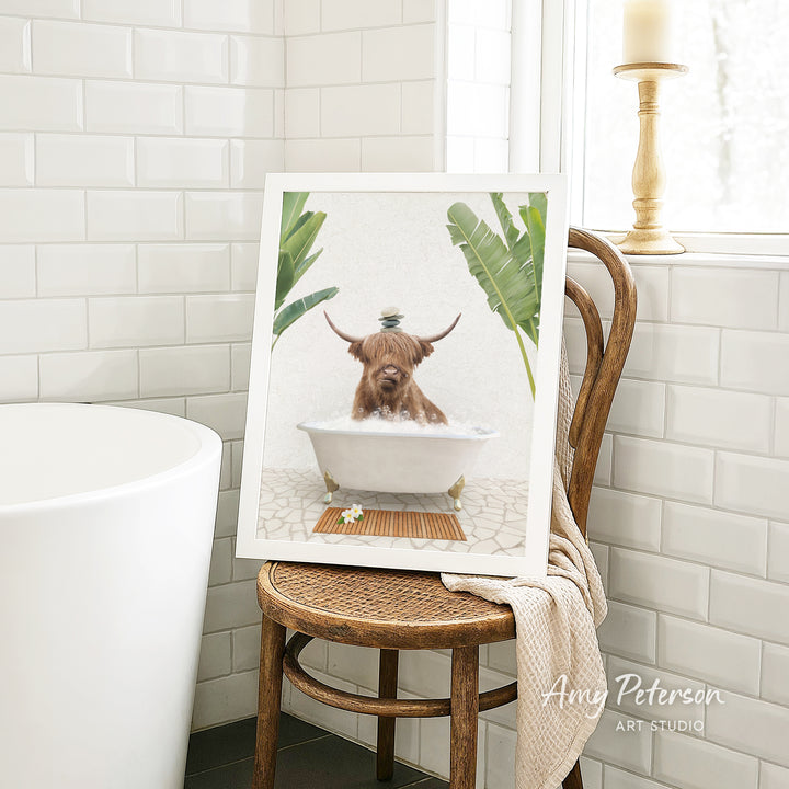 A framed image of a brown cow sitting in a white bathtub, surrounded by green plants, is displayed on a wooden chair in a bathroom setting.