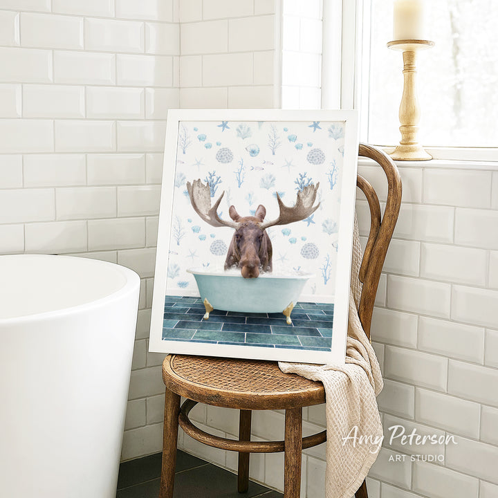 A framed image of a moose sitting in a bathtub, surrounded by blue and white floral patterns, is displayed on a wooden chair in a bathroom setting.