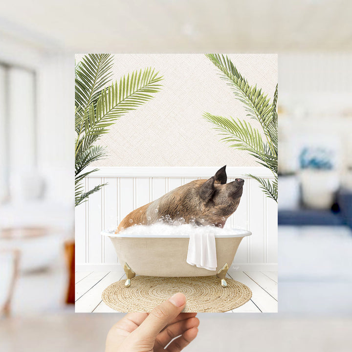 A hand holds up a framed image depicting a pig sitting in a bathtub filled with bubbles, surrounded by palm leaves.