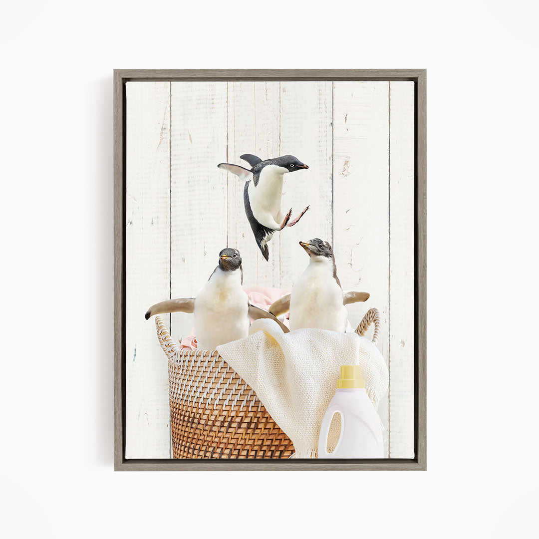 A framed photograph of three penguins sitting in a woven basket, with a bottle of detergent and a towel visible in the background.