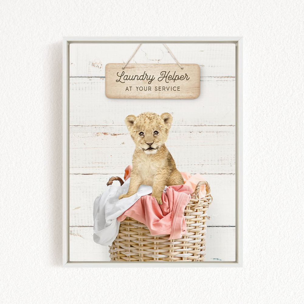 A young lion cub sitting in a wicker basket filled with clothes, with a wooden sign above that reads "Laundry Helper at your service".