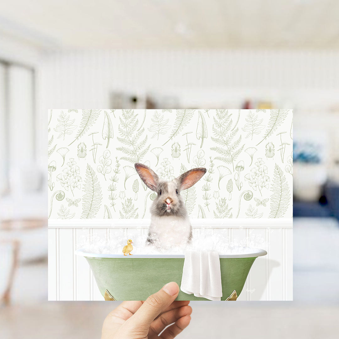 A hand holding a framed print of a rabbit sitting in a bathtub filled with water, surrounded by a green and white floral pattern.