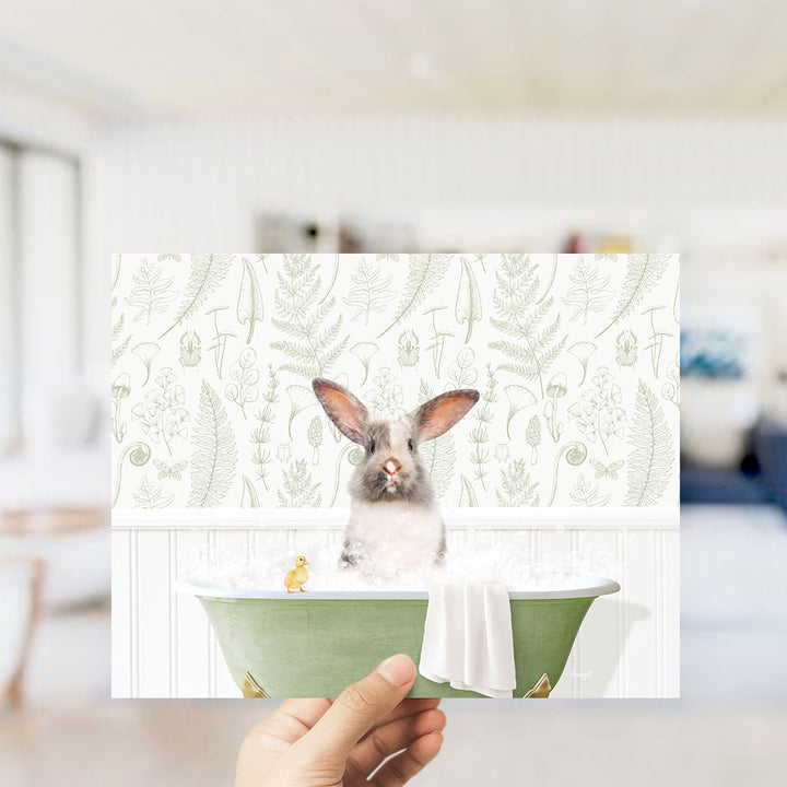 A hand holding a framed print of a rabbit sitting in a bathtub filled with water, surrounded by a green and white floral pattern.