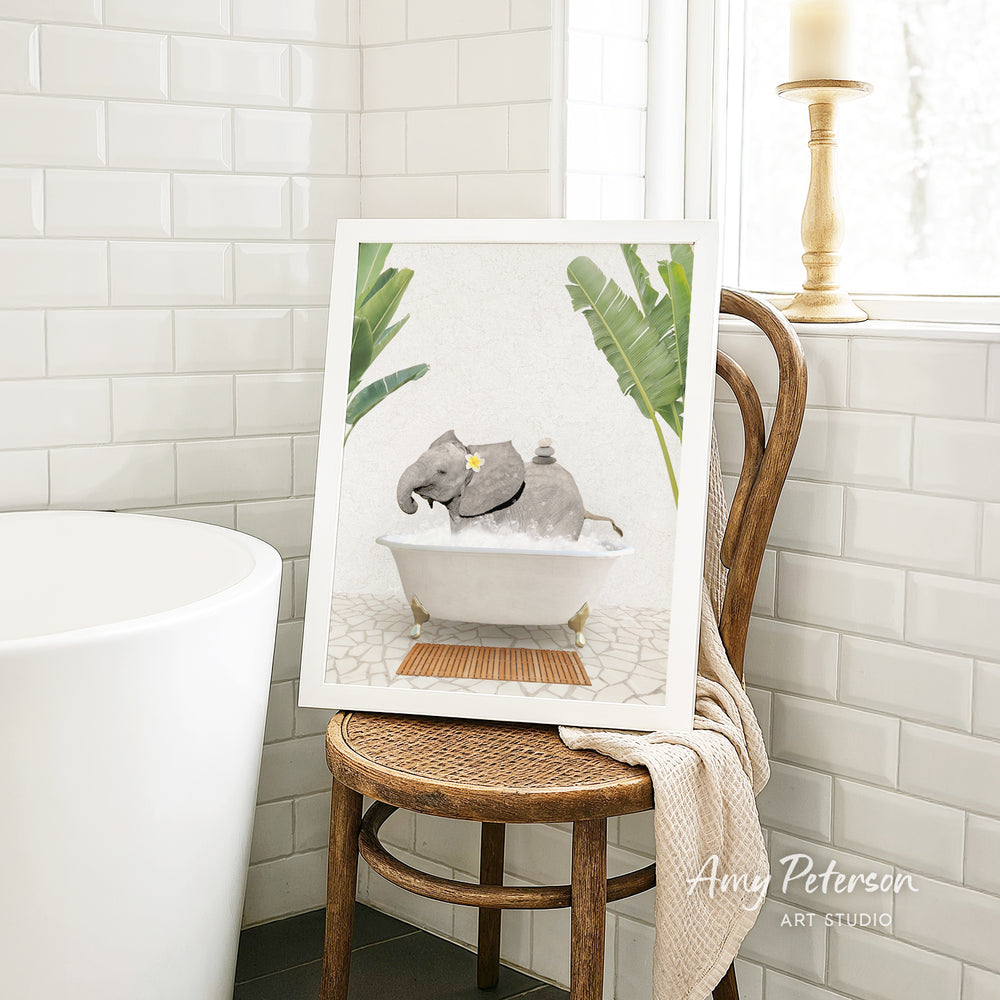A framed image of a gray elephant sitting in a bathtub, surrounded by green leaves, is displayed on a wooden chair in a bathroom setting.