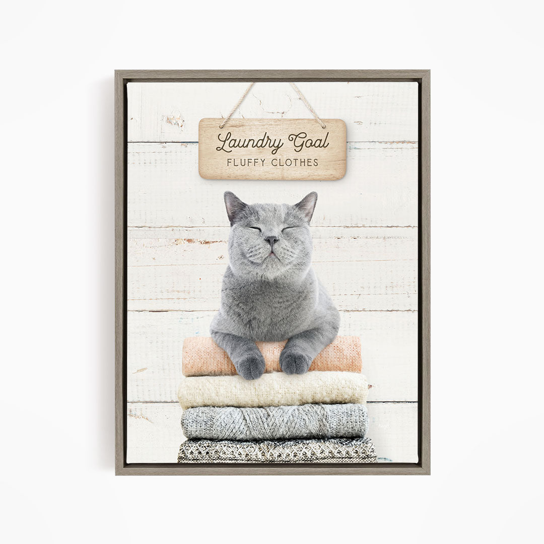 A gray cat sitting on a stack of folded clothes, with a wooden sign above that says "Laundry Goal Fluffy Clothes".