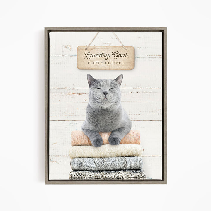 A gray cat sitting on a stack of folded clothes, with a wooden sign above that says "Laundry Goal Fluffy Clothes".