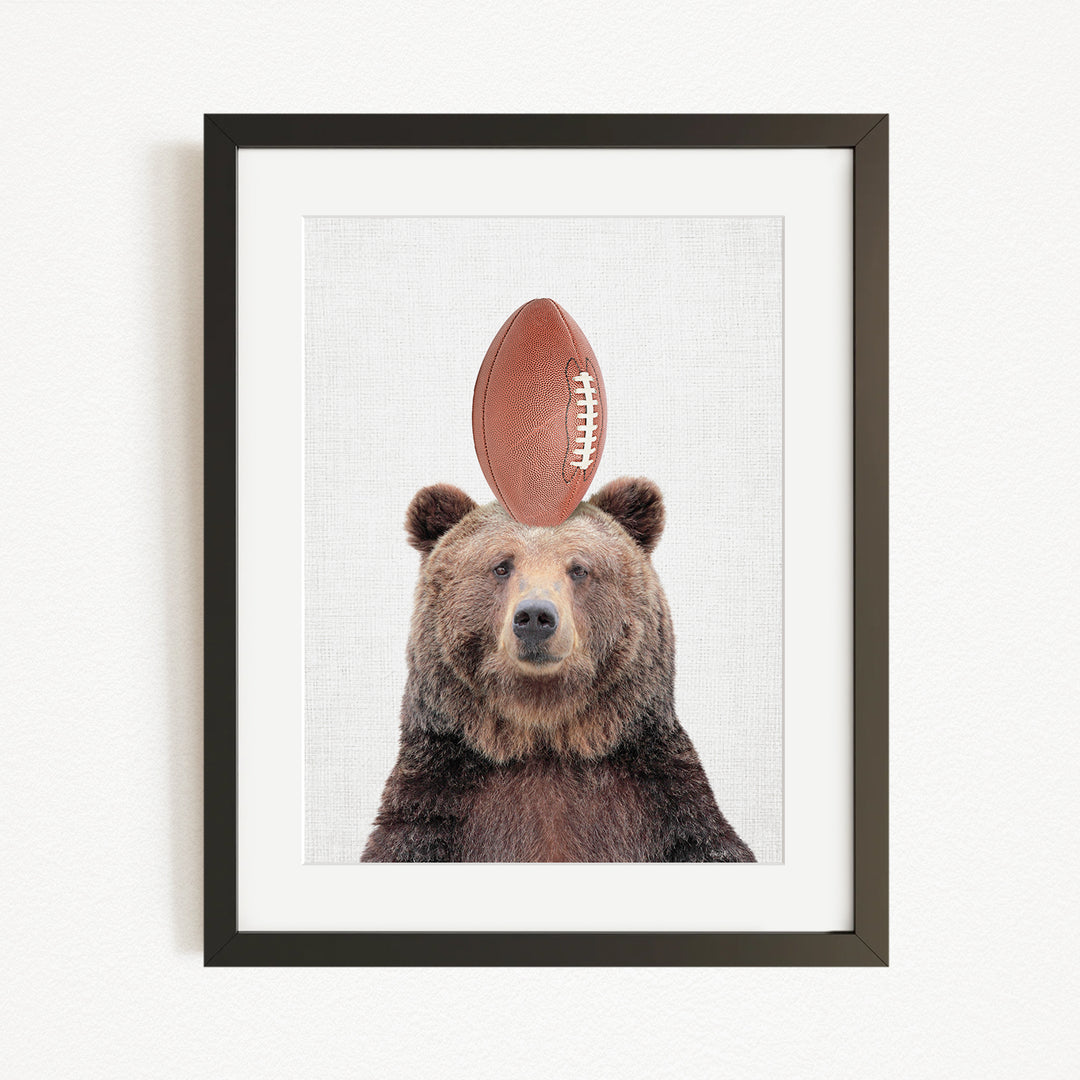 A framed image of a brown bear with a football on its head.