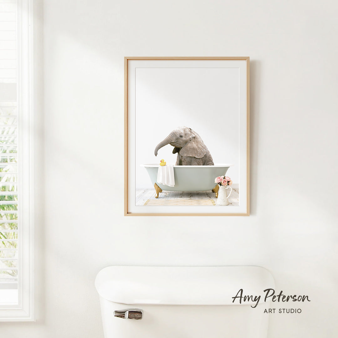 A framed image of an elephant sitting in a bathtub, with a vase of flowers and a towel nearby, is hanging on a white wall.