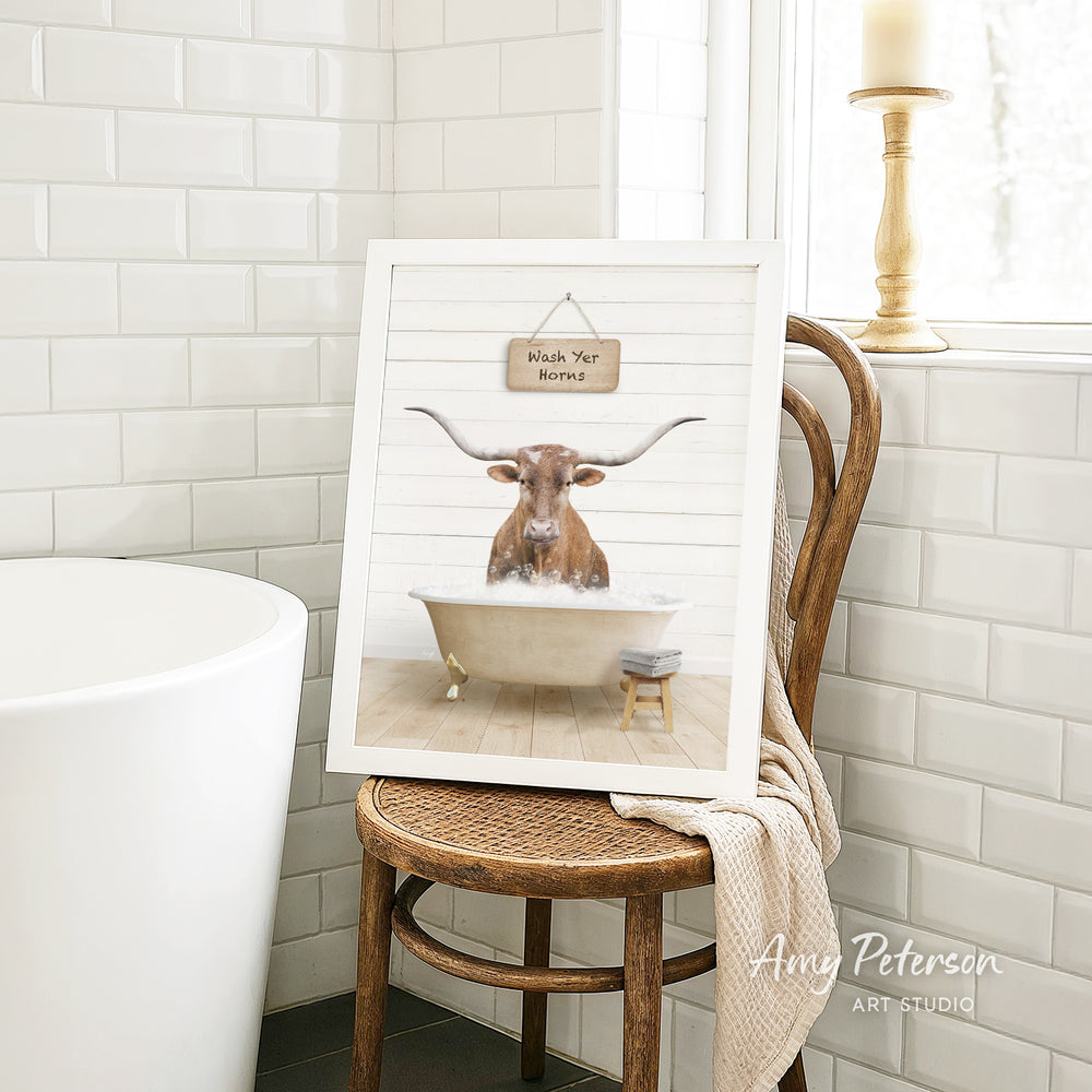 A framed print of a bull with horns sitting in a bathtub, accompanied by a wooden chair and a towel in a bathroom setting.