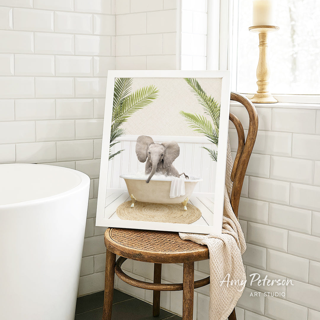 A framed image of a baby elephant sitting in a bathtub, surrounded by palm leaves, is displayed on a wooden chair in a bathroom setting.