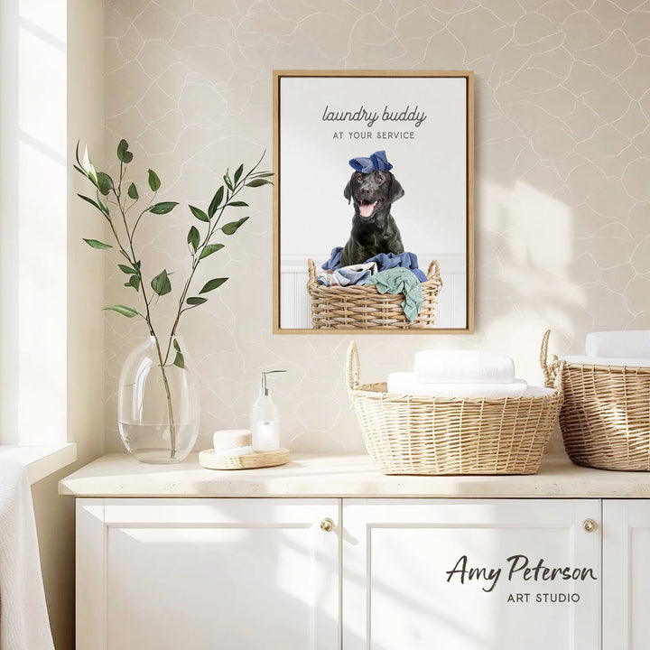 a framed photo of a black dog sitting in a wicker basket, with the caption "laundry buddy at your service" above it. The photo is displayed on a wall above a white cabinet, which has a vase of green leaves and a bottle of soap on it.