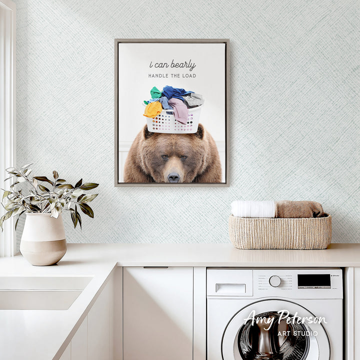 a framed poster of a bear with a basket of laundry on its head, hanging on a wall above a white countertop with a potted plant and a basket of towels.