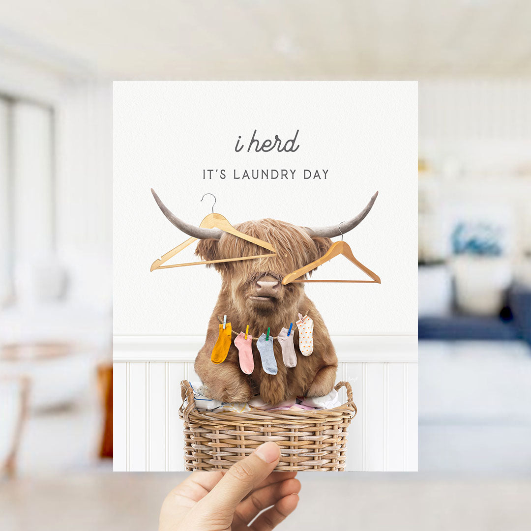 A hand holds up a card featuring a brown cow with horns wearing clothes and socks, sitting in a wicker basket with clothes hanging on hangers.
