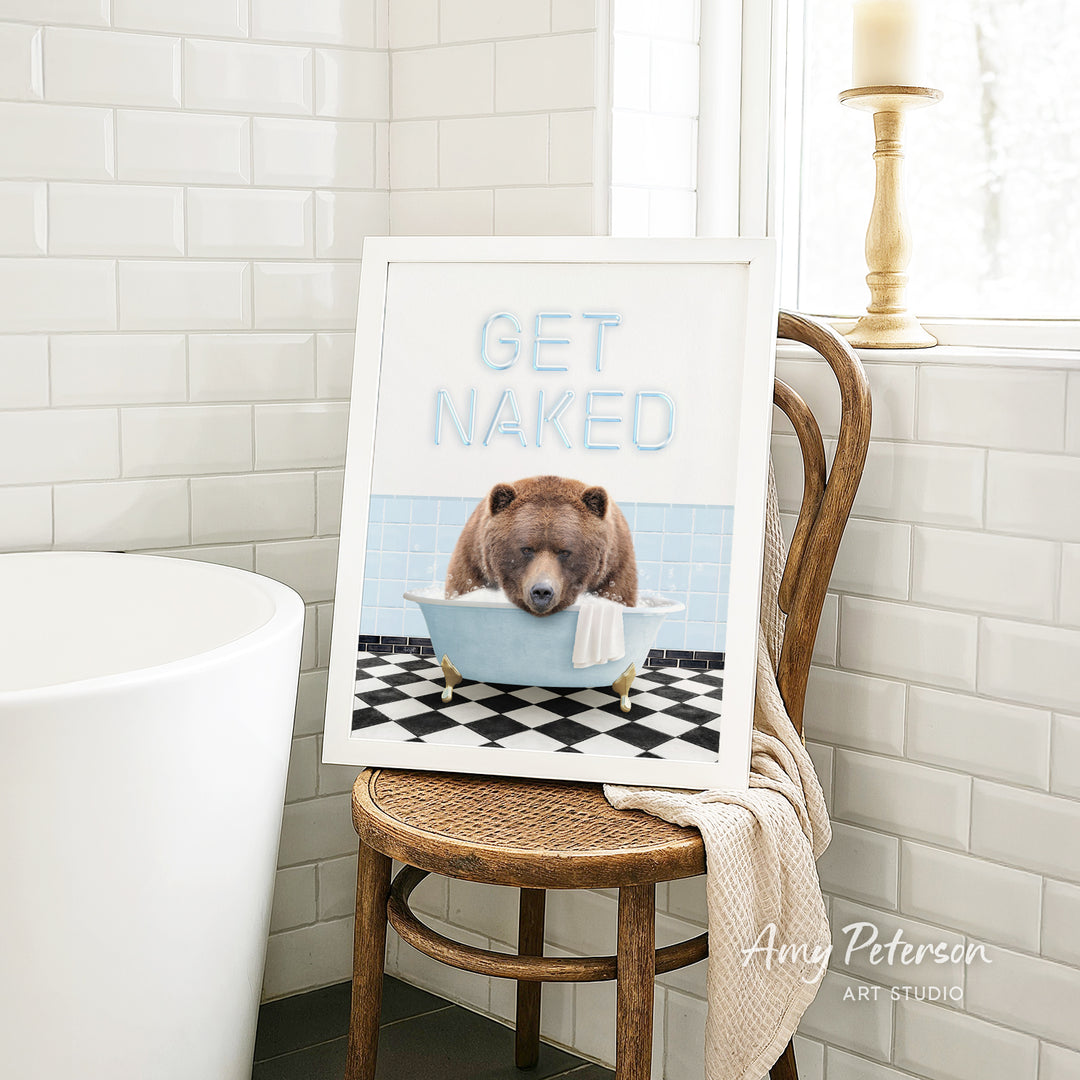 A framed print featuring a bear sitting in a bathtub with the text "GET NAKED" on it, placed on a wooden chair in a bathroom setting.