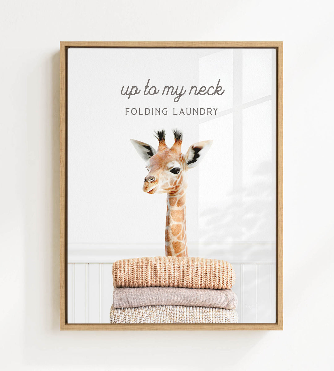 A framed image of a giraffe standing on top of a stack of folded clothes, with the text "up to my neck" and "FOLDING LAUNDRY" written above the image.