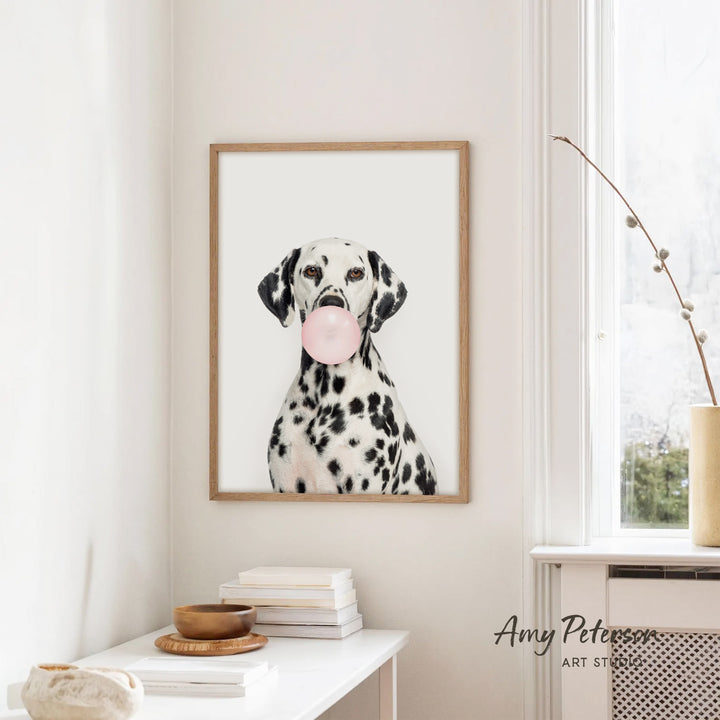 A framed photograph of a Dalmatian dog holding a pink bubble gum bubble is hanging on a white wall in a room with a white table and a window.