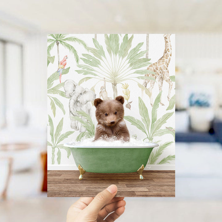 A hand holds up a framed print featuring a small brown bear sitting in a green bathtub, surrounded by a tropical jungle scene with various animals and plants.