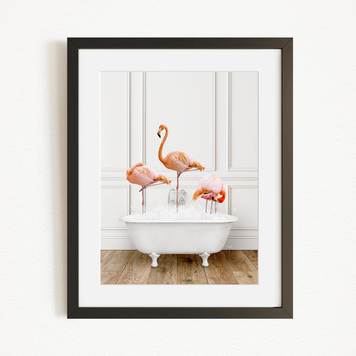 A framed image depicting three flamingos standing in a white bathtub, with a white wall and wooden floor in the background.