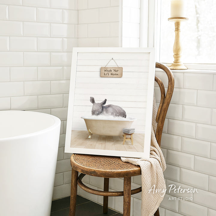 A framed print of a gray rabbit sitting in a bathtub is displayed on a wooden chair in a bathroom setting.