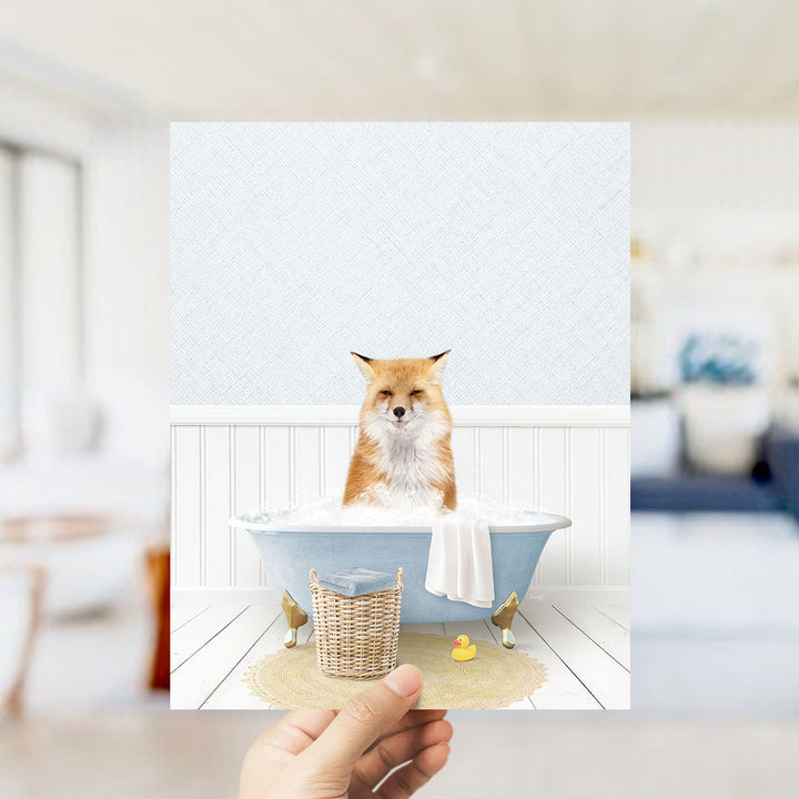 A hand holds up a framed image depicting a fox sitting in a bathtub filled with bubbles, surrounded by a yellow rubber duck and a white towel.